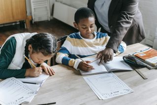 High angle of crop unrecognizable black female teacher explaining task to focused little schoolboy sitting at desk near attentive Asian classmate writing in notebook